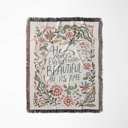 A rectangular woven throw blanket with fringe on all sides, featuring a cream background and a central quote that reads 'He Makes Everything Beautiful In Its Time' in dark green lettering. The quote is surrounded by a decorative border of hand-drawn style flowers and leaves in muted pink, teal, olive green, and brown tones.