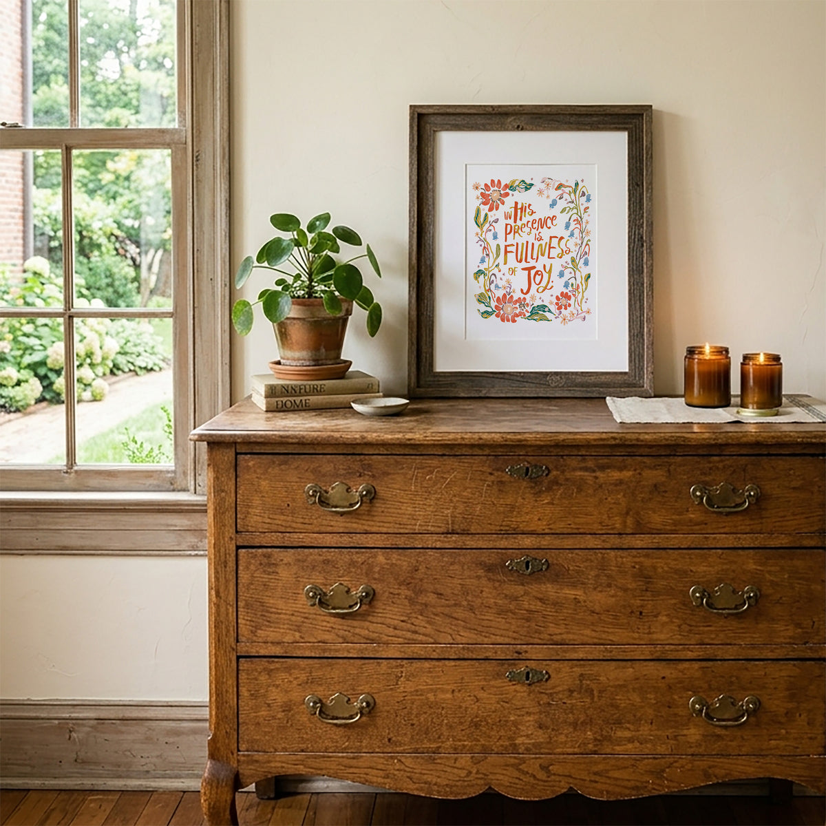A framed art print with colorful floral illustrations and the words 'This happens 30 times to joy!' displayed on a rustic wooden chest of drawers next to a window.