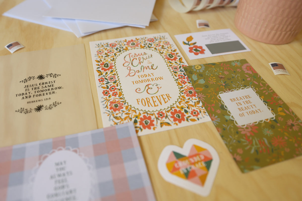 A collection of floral and typographic greeting cards and small stickers arranged on a light wood tabletop. The cards feature colorful botanical borders, inspirational quotes, and pastel plaid accents.