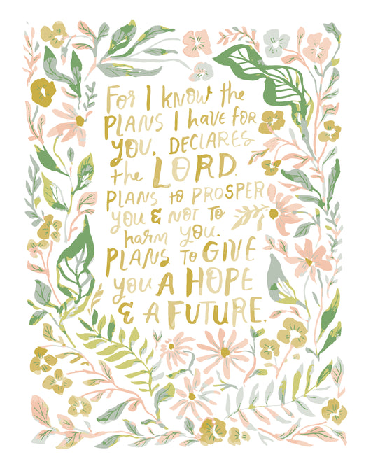 A square art print featuring a hand-lettered inspirational quote in gold text surrounded by a border of delicate watercolor-style flowers and leaves in soft green, pink, and yellow on a white background.
