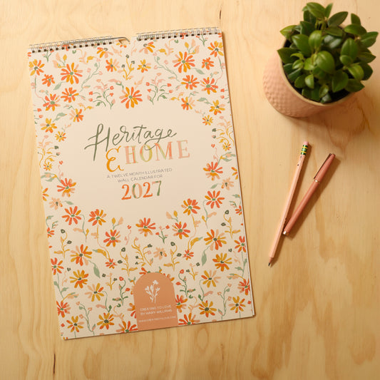 A 2027 spiral-bound wall calendar titled 'Heritage & Home' with a floral pattern, displayed on a light wood tabletop next to a small potted plant and two pink pens.