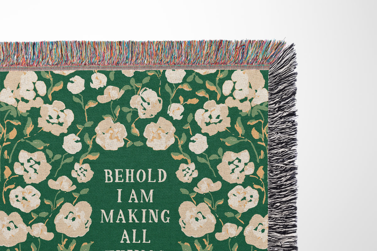 A close-up of a woven throw blanket corner showing a dark green background with cream floral pattern, multicolor fringe, and the text 'BEHOLD I AM MAKING ALL THESE' in white.