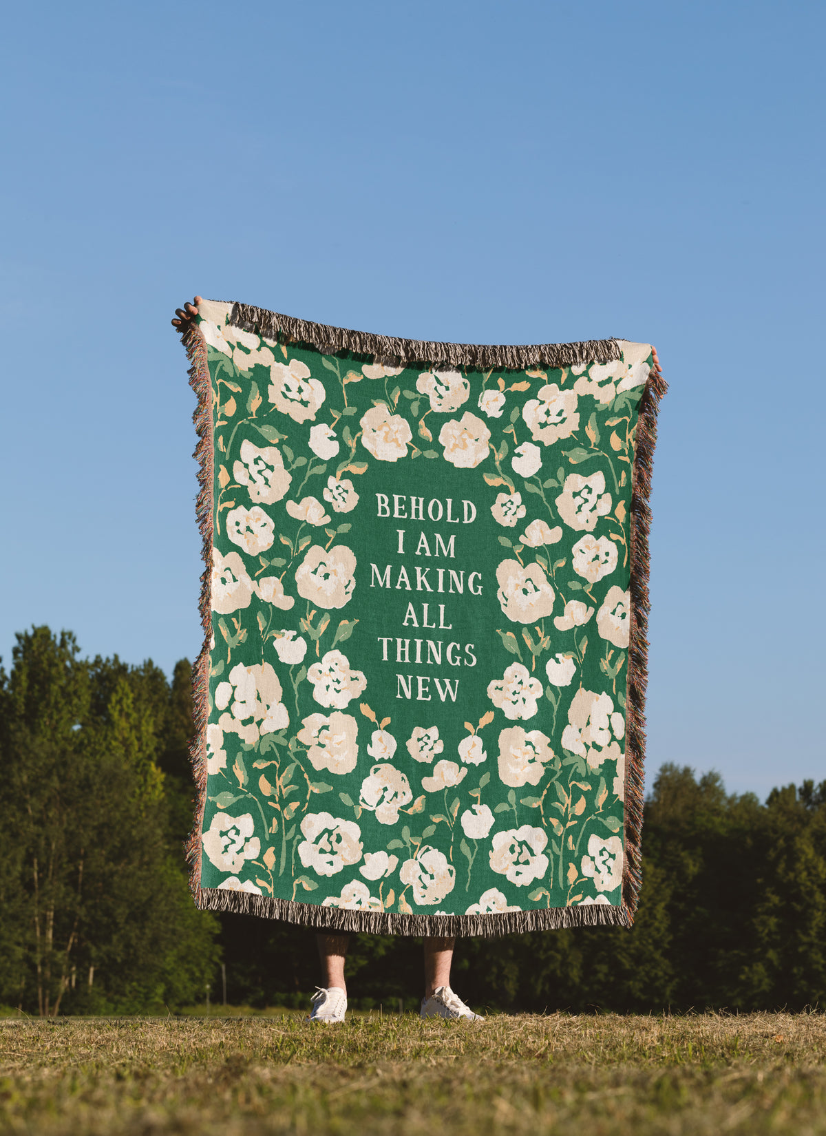 A person standing outdoors in a grassy field under a blue sky holds up a large woven throw blanket. The blanket is green with a cream floral pattern and the text 'BEHOLD I AM MAKING ALL THINGS NEW' in the center.