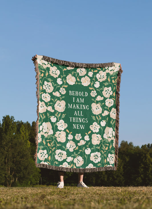 A person standing outdoors in a grassy field under a blue sky holds up a large woven throw blanket. The blanket is green with a cream floral pattern and the text 'BEHOLD I AM MAKING ALL THINGS NEW' in the center.