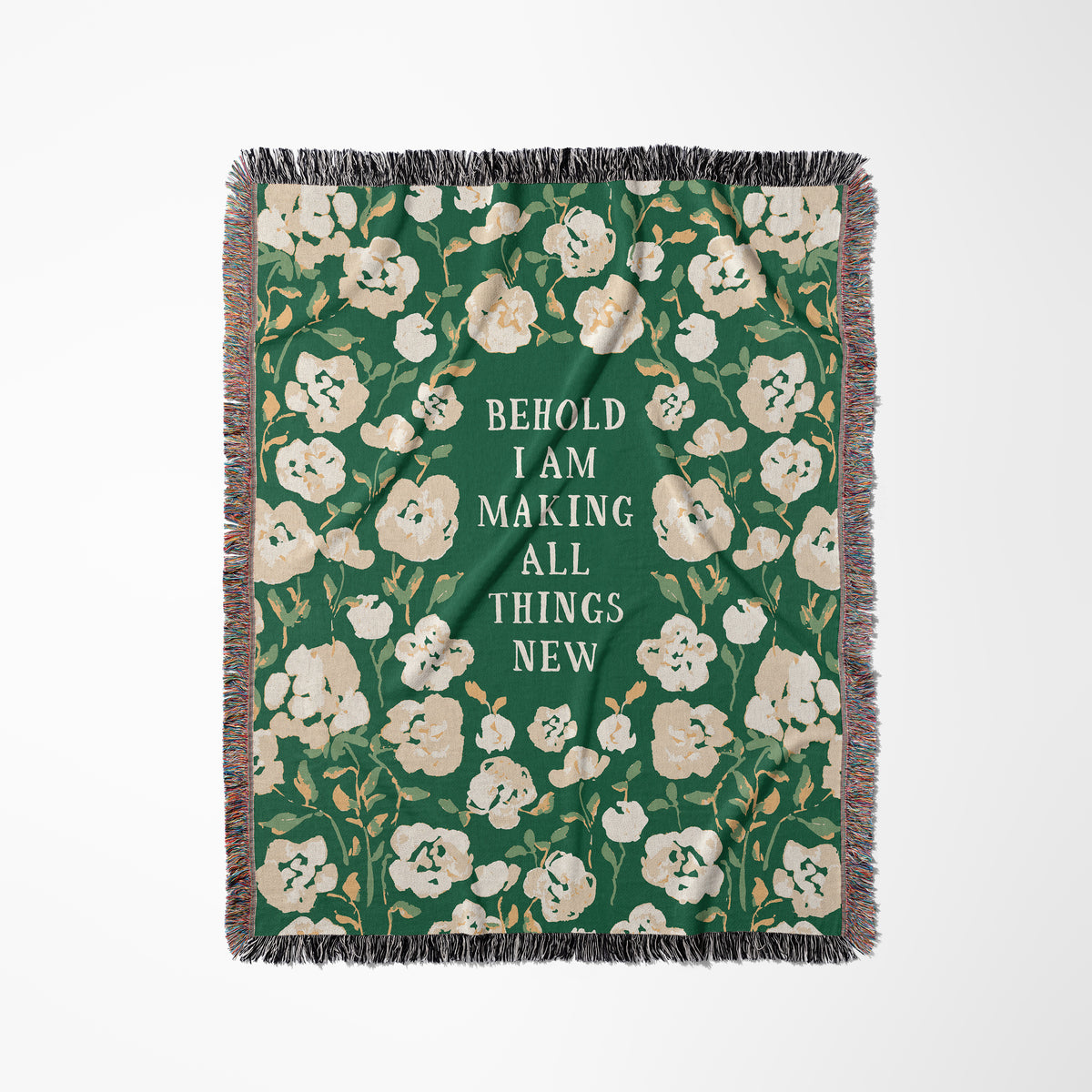 A rectangular woven throw blanket with fringe on all sides, featuring a dark green background with cream and beige floral motifs and centered white text that reads, 'BEHOLD I AM MAKING ALL THINGS NEW.'.