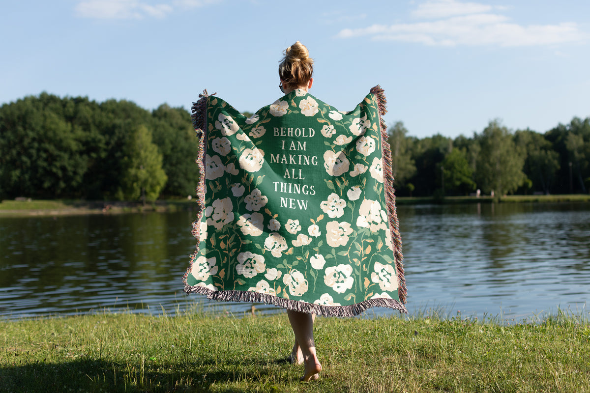A woman stands on a grassy lakeside with her back to the camera, holding up a large green tapestry with white floral patterns and the text 'BEHOLD I AM MAKING ALL THINGS NEW'.