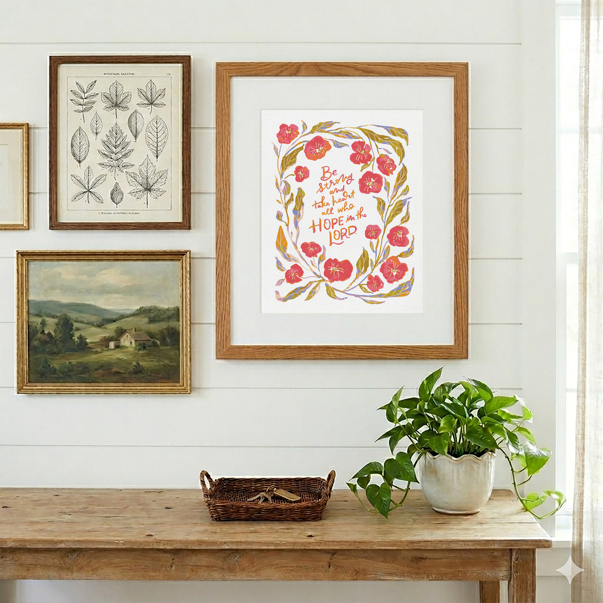 A framed art print featuring a floral wreath and the quote 'Be not afraid to have the hope of your LORD' in red and orange lettering, styled on a white paneled wall above a rustic wooden table with a potted green plant.