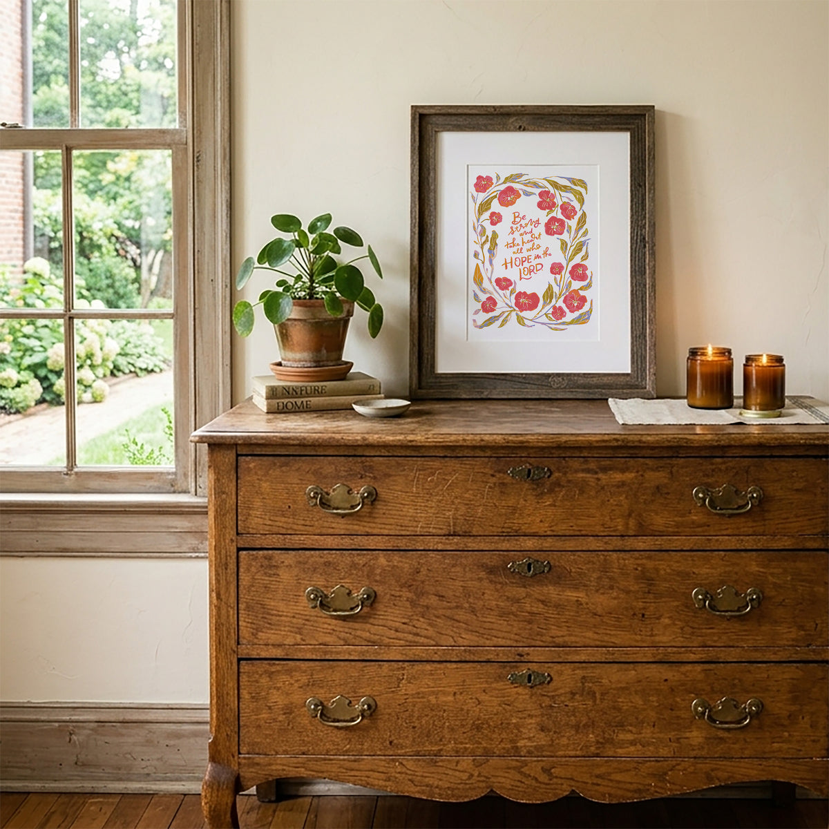 A framed art print featuring a watercolor floral wreath with the words 'Love is patient, love is kind' displayed on a rustic wooden dresser next to a window, with a potted plant and candles.