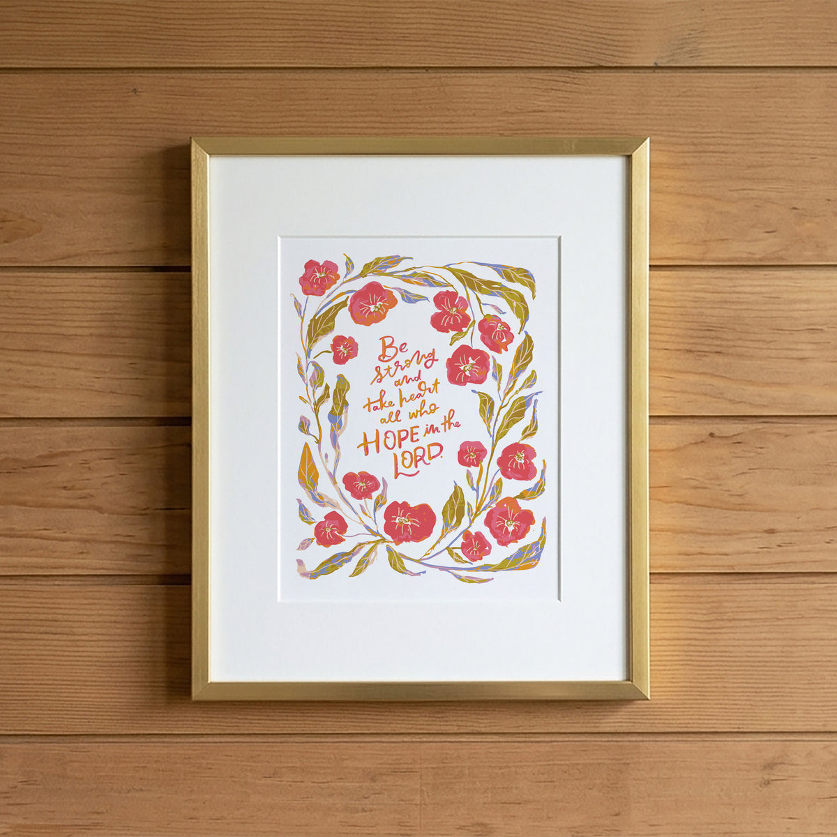 A framed art print featuring a floral wreath illustration with the hand-lettered Bible verse 'Be joyful and hope in the Lord' on a white mat, set in a gold frame against a rustic wooden plank background.