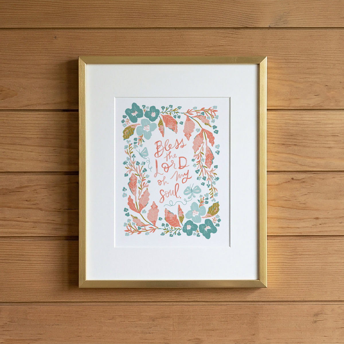 A framed art print featuring the hand-lettered quote 'Blessed is my soul' surrounded by a watercolor floral wreath in teal, coral, and sage green, displayed in a gold frame against a light brown wooden plank wall.