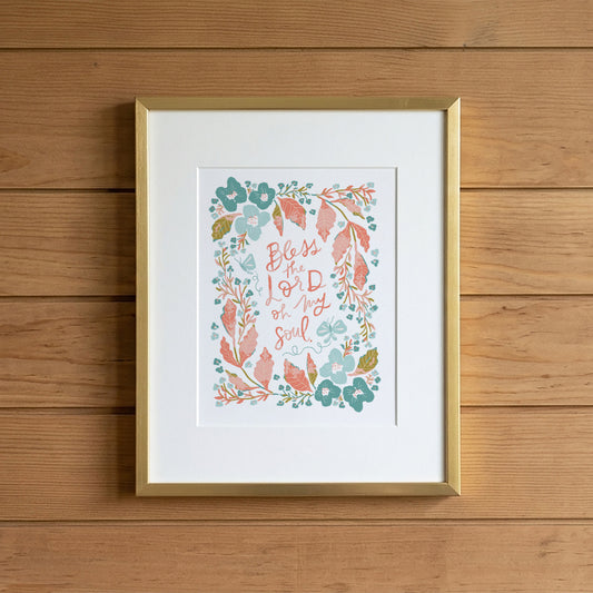 A framed art print featuring the hand-lettered quote 'Blessed is my soul' surrounded by a watercolor floral wreath in teal, coral, and sage green, displayed in a gold frame against a light brown wooden plank wall.