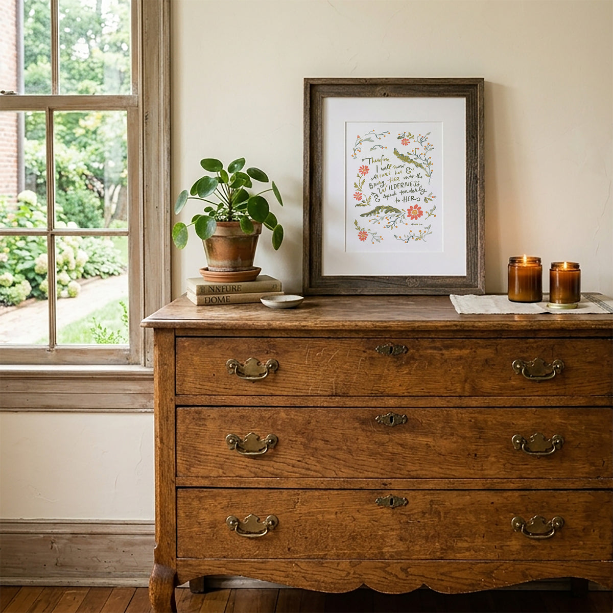 A framed botanical art print with the quote 'The best things in life are free' displayed on a rustic wooden dresser next to a window, with a potted plant and candles on top.