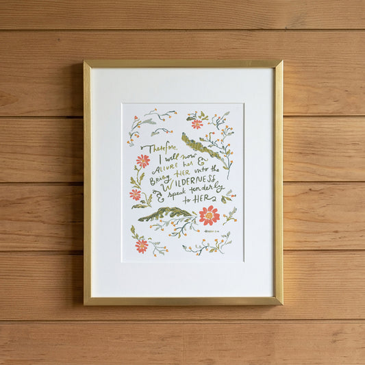 A framed art print featuring a botanical illustration with a handwritten quote, displayed on a light wood plank wall. The print has a white mat, delicate watercolor-style flowers and leaves in green, orange, and pink, and is enclosed in a thin gold frame.