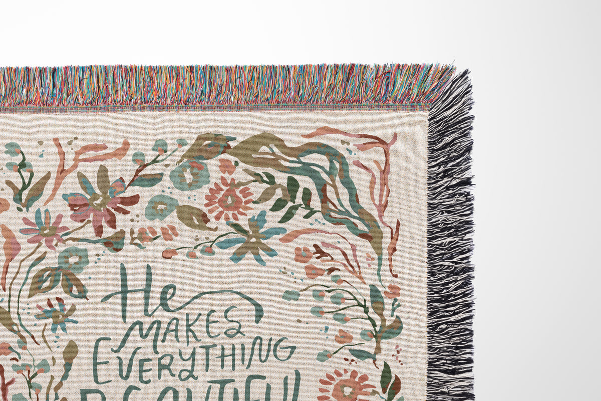A close-up of a woven throw blanket corner featuring multicolor fringe, a cream linen-textured base, and a floral border with the phrase 'He makes everything beautiful' in dark teal script.