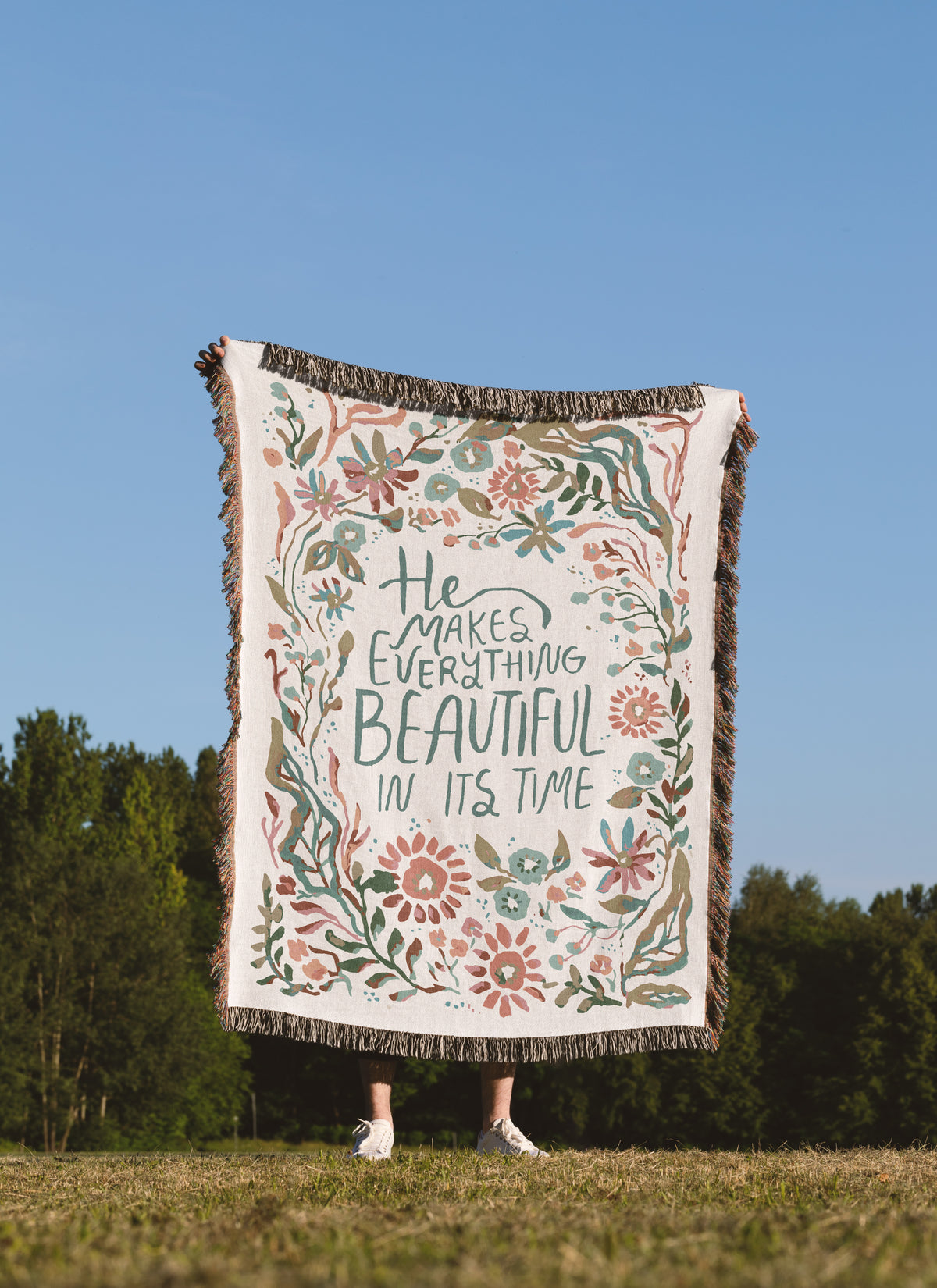 A person standing outdoors in a grassy field under a blue sky holds up a large woven throw blanket. The blanket has a cream background with a floral border in muted pink, green, and blue, and the phrase 'He Makes Everything Beautiful In Its Time' printed in the center.