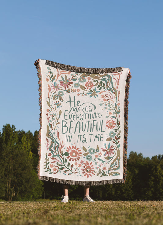 A person standing outdoors in a grassy field under a blue sky holds up a large woven throw blanket. The blanket has a cream background with a floral border in muted pink, green, and blue, and the phrase 'He Makes Everything Beautiful In Its Time' printed in the center.