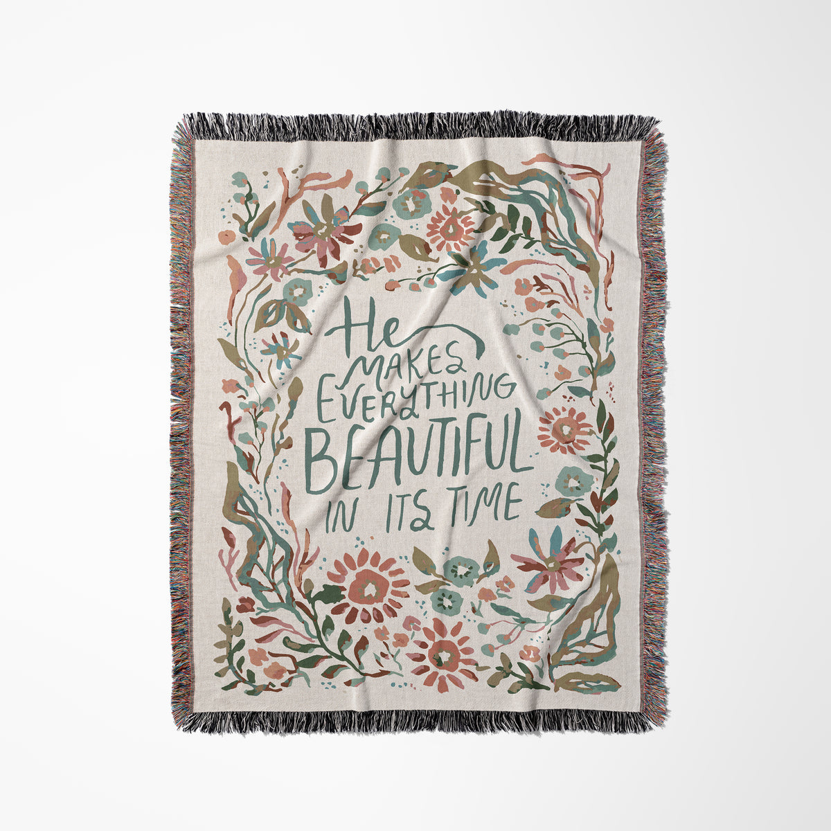 A rectangular woven throw blanket with fringe on all sides, featuring a cream background and a central quote that reads 'He Makes Everything Beautiful In Its Time' in dark green lettering. The quote is surrounded by a decorative border of hand-drawn style flowers and leaves in muted pink, teal, olive green, and brown tones.