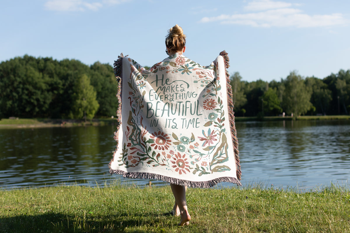 A woman stands on a grassy lakeside with trees and blue sky in the background, holding up a large cream throw blanket with fringe and a floral print. The back of the blanket features the quote, 'He makes everything beautiful in its time' in black script.