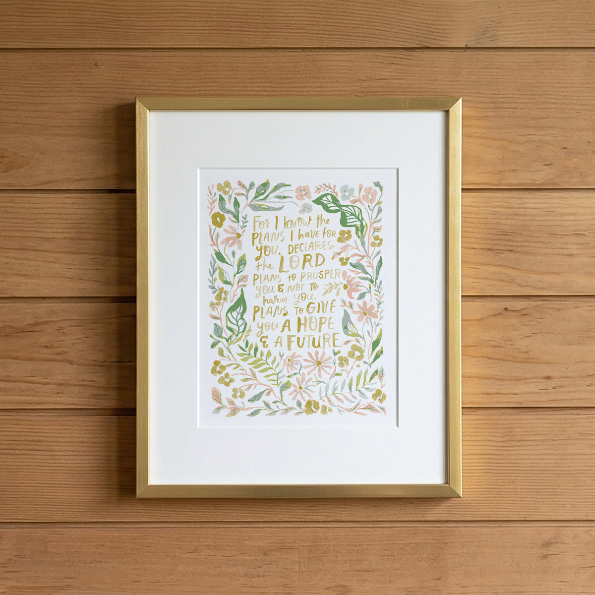 Framed floral wall art with a gold frame and white mat, featuring a central botanical illustration with gold lettering, displayed against a warm wood plank wall.