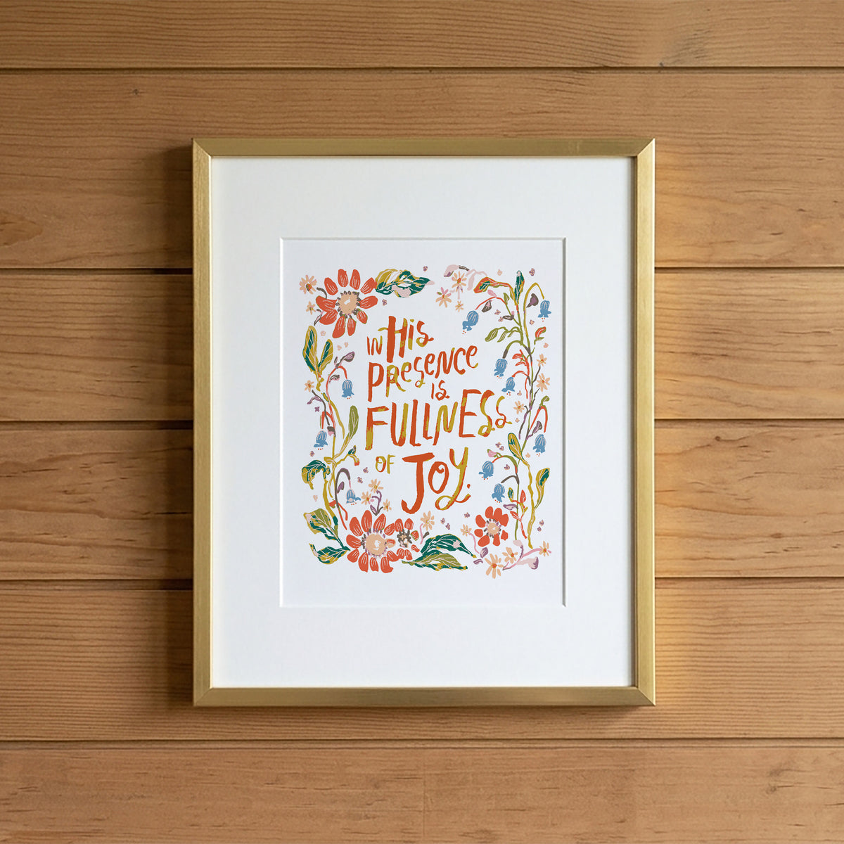 Framed wall art with a gold frame and white mat, featuring a colorful floral illustration and the text 'In His Presence is Fullness of Joy,' displayed against a wood plank wall.