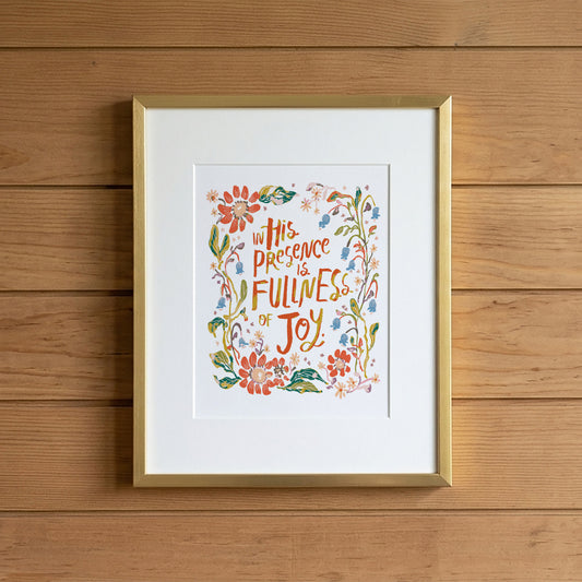 Framed wall art with a gold frame and white mat, featuring a colorful floral illustration and the text 'In His Presence is Fullness of Joy,' displayed against a wood plank wall.