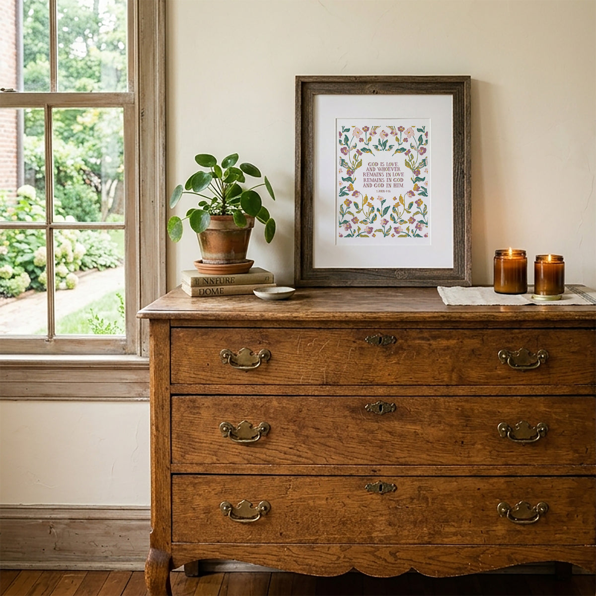 A framed art print featuring a floral border and a quote is displayed on a rustic wooden dresser next to a window, with a potted plant and candles on top.