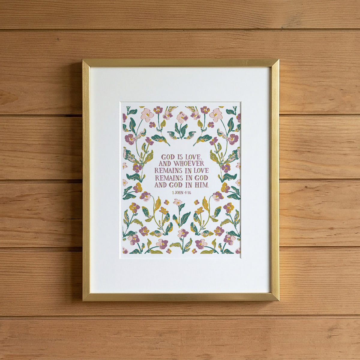 A framed floral Bible verse print featuring a watercolor-style botanical border and the text 'GOD IS LOVE, AND WHOEVER REMAINS IN LOVE REMAINS IN GOD AND GOD IN HIM 1 JAMES 2:15-16' on a white mat, set in a gold frame against a light wood plank wall.