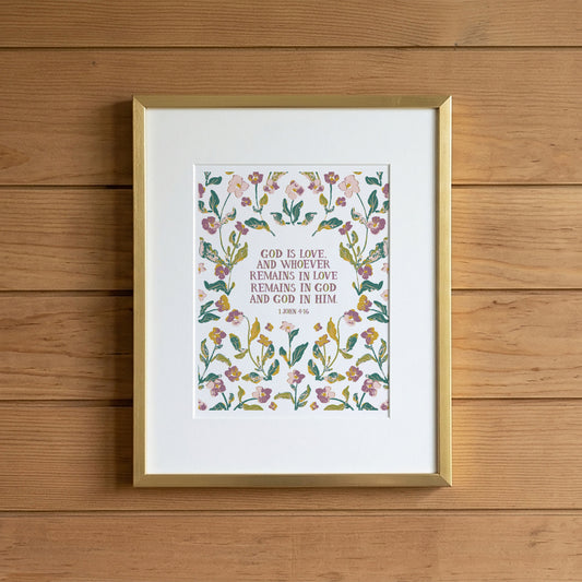 A framed floral Bible verse print featuring a watercolor-style botanical border and the text 'GOD IS LOVE, AND WHOEVER REMAINS IN LOVE REMAINS IN GOD AND GOD IN HIM 1 JAMES 2:15-16' on a white mat, set in a gold frame against a light wood plank wall.