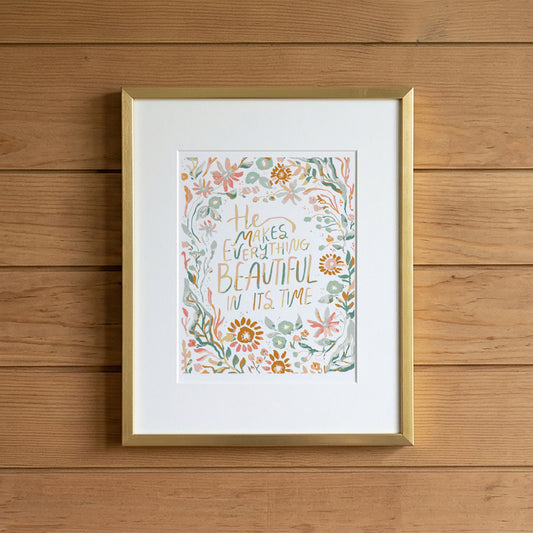 A framed art print featuring a floral border and the hand-lettered quote 'He makes everything beautiful in its time,' displayed against a light brown wooden plank wall.
