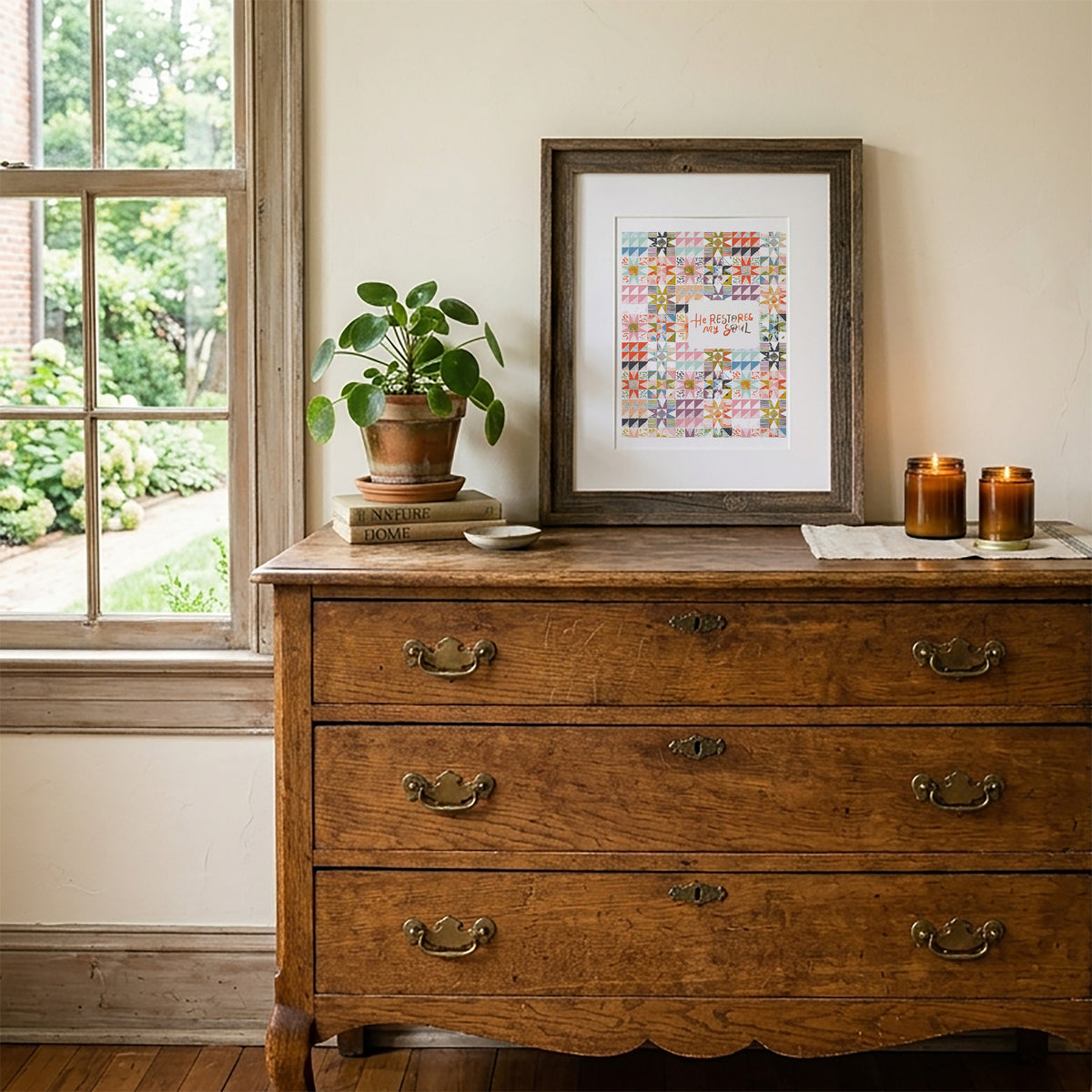A framed art print with a colorful patchwork design in a dark wood frame, styled on top of a vintage wooden chest of drawers next to a window.