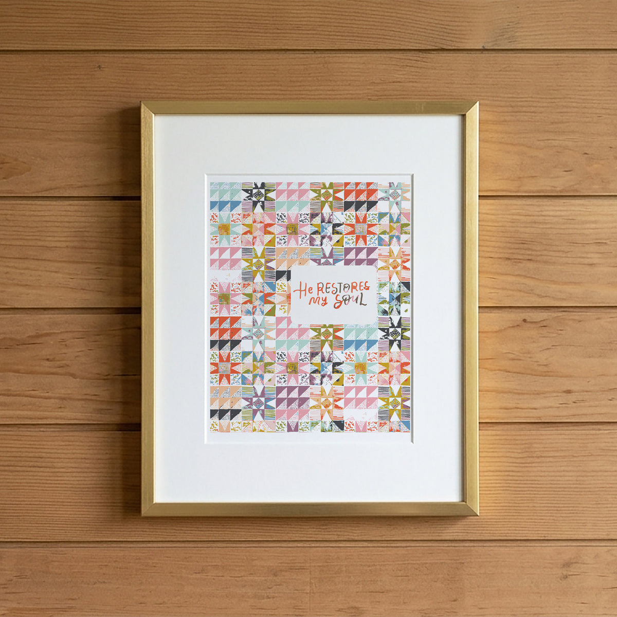 A framed art print featuring a colorful quilt pattern with the text 'He Rests My Soul' in the center, displayed on a light wood plank background.