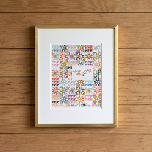 A framed art print featuring a colorful quilt pattern with the text 'He Rests My Soul' in the center, displayed on a light wood plank background.