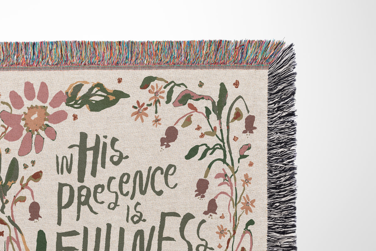 A close-up of a woven textile with fringe, featuring floral illustrations and the phrase 'in His Presence is Fullness' on a light neutral background.