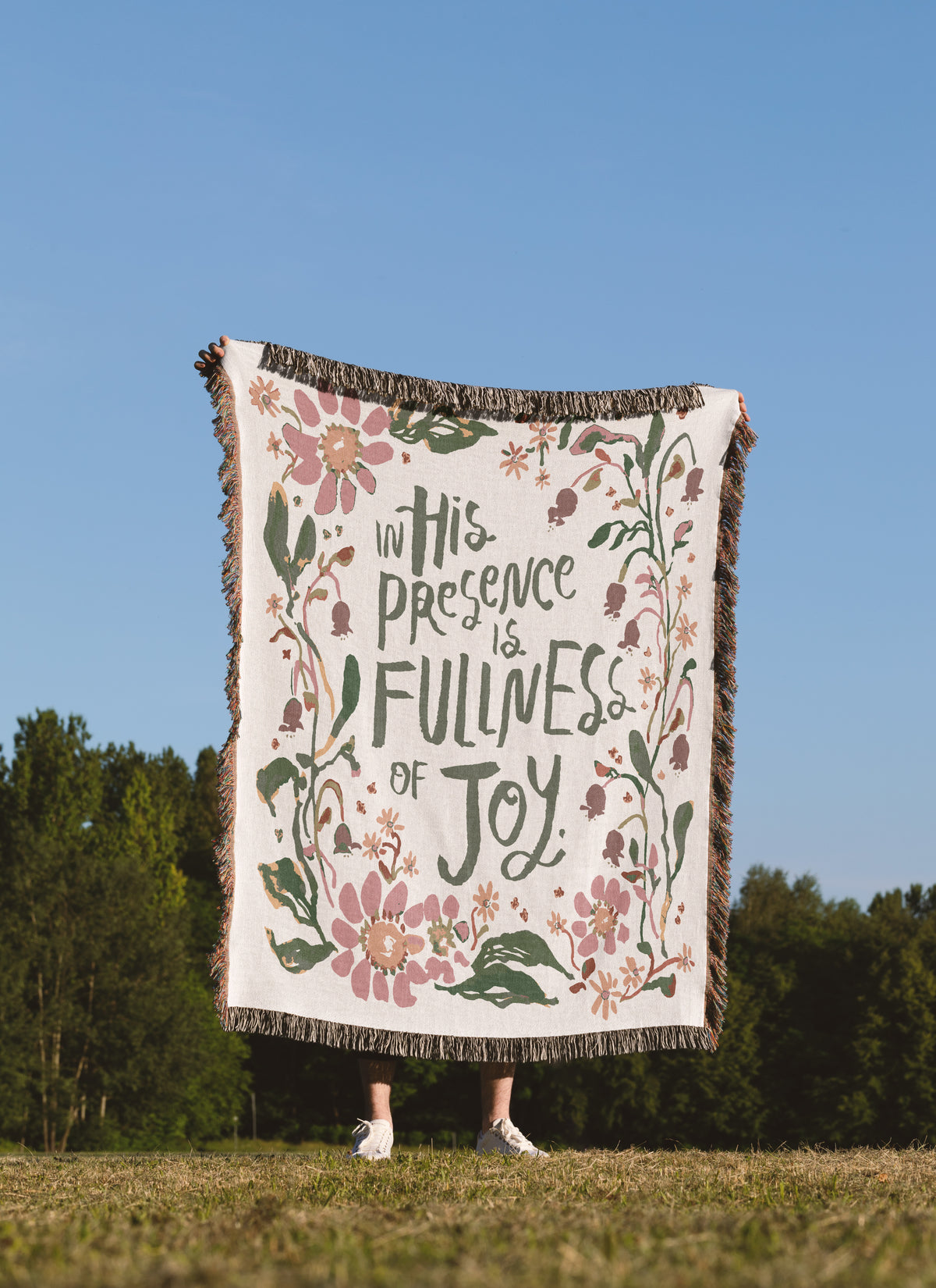 A person standing outdoors in a grassy field under a clear blue sky holds up a large woven blanket or throw. The blanket has a cream background with pink floral illustrations, green leaves, and dark lettering that reads, 'in His Presence is Fullness of Joy.