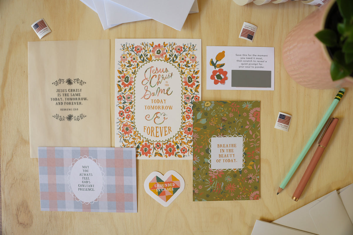 A styled flat-lay of floral and typographic greeting cards, envelopes, stickers, pencils, and paper on a light wood surface. The central card reads 'Love Never Ends Today Tomorrow Ever', surrounded by coordinating stationery items.