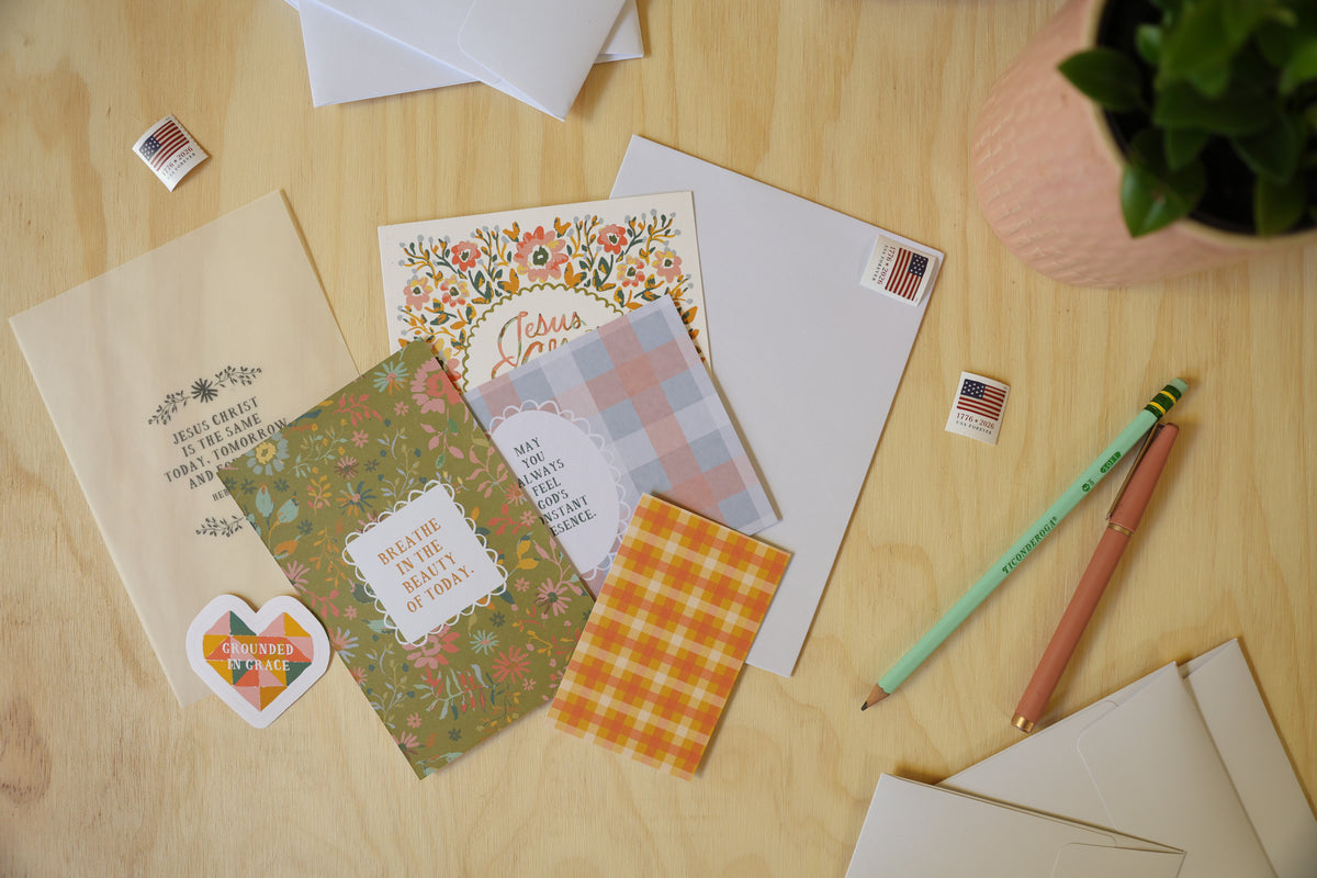 A styled flat-lay of handmade greeting cards, envelopes, and small American flag stickers on a light wood tabletop, with a green plant in a pink pot at the top right.