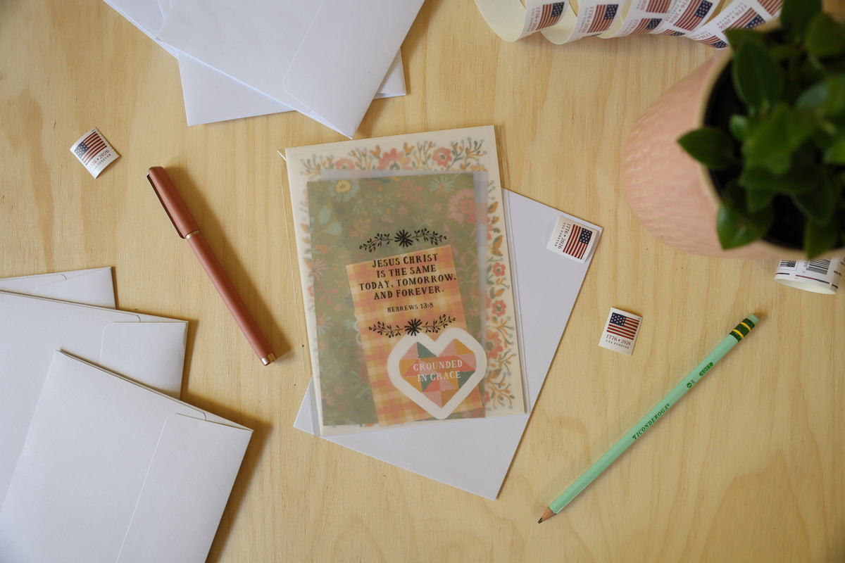 A flat-lay view of a handmade greeting card with a floral border and a heart-shaped 'Love Never Fades' sticker, displayed on a light wood desk alongside white envelopes, a rose gold pen, a green pencil, and a potted plant.