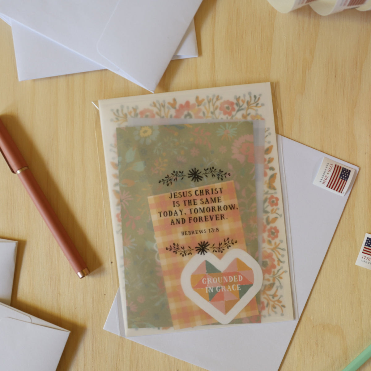 A styled flat-lay of a personalized Bible verse card with a floral border and a heart reading 'Ordained Vocation,' surrounded by white envelopes, small American flag stickers, a rose gold pen, and a mint green pencil on a light wood desk.