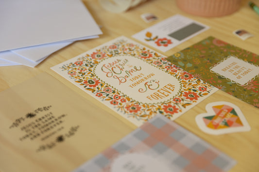 A close-up of a floral wedding invitation card with the words 'Love You Today Tomorrow & Forever' in an oval frame, surrounded by red and orange flowers. Other matching stationery items are partially visible on a light wood surface.