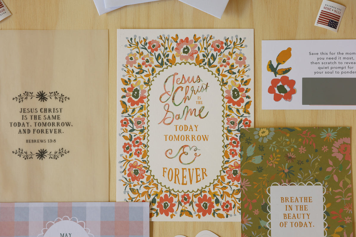 A styled flat-lay of floral greeting cards and stationery on a light wood surface. The main card in the center reads “Jesus Christ is in the Balm Today Tomorrow & Forever” with a multicolor floral border.