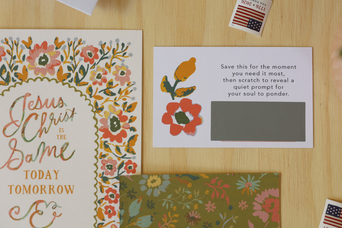 A flat-lay view of a stationery set on a light wood background, featuring a floral 'Jesus Christ Is The Same' card, a matching prompt card with a gray scratch-off box, and a green floral envelope.