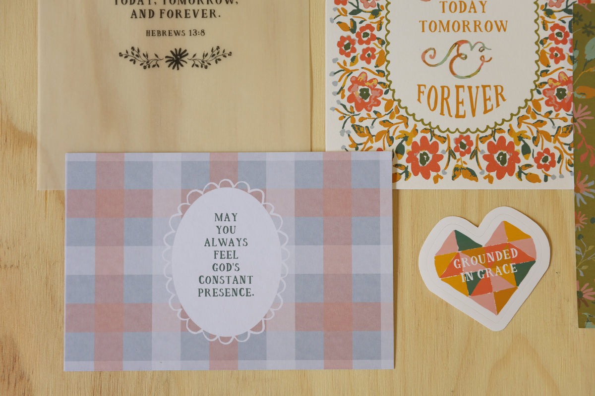 A styled flat-lay of several inspirational cards on a light wood background. The cards feature various designs: a beige card with black text, a white card with a colorful floral border and orange text, a blue and pink gingham card with a white oval label, and a small heart-shaped card with a geometric multicolor pattern.
