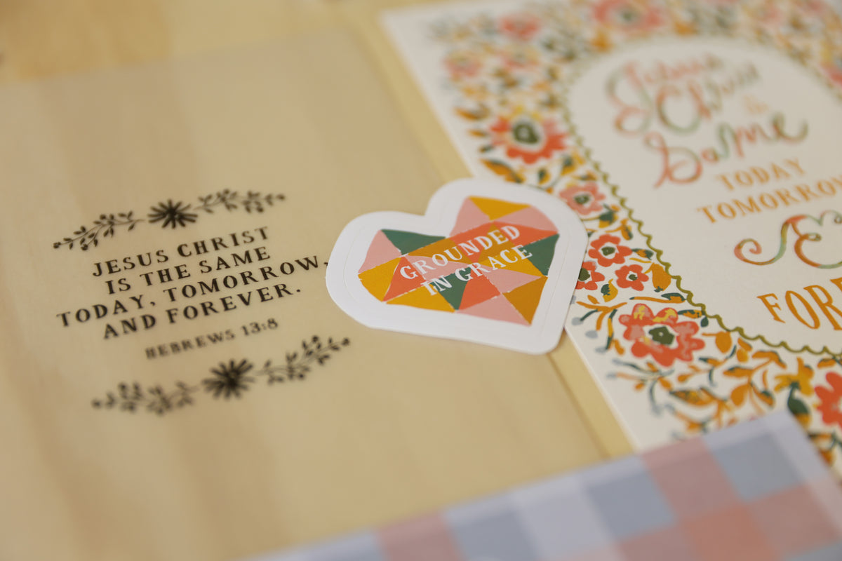 A heart-shaped sticker with a colorful geometric pattern and the words 'Grounded in Grace' placed on top of cream-colored stationery with floral and typographic designs.