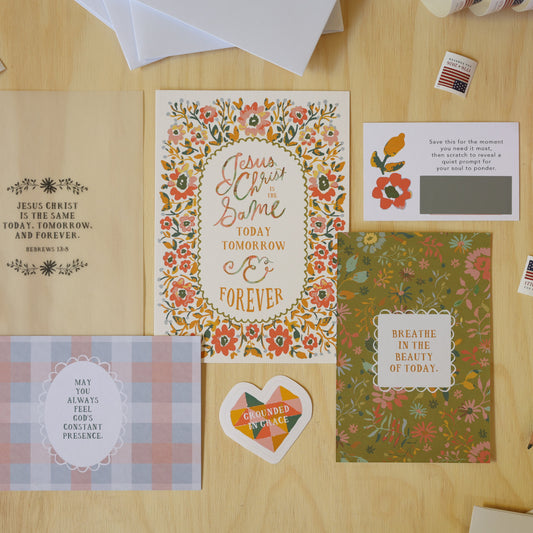 A styled flat-lay of handmade greeting cards and envelopes on a light wood desk, surrounded by pencils, a plant, and small stickers.