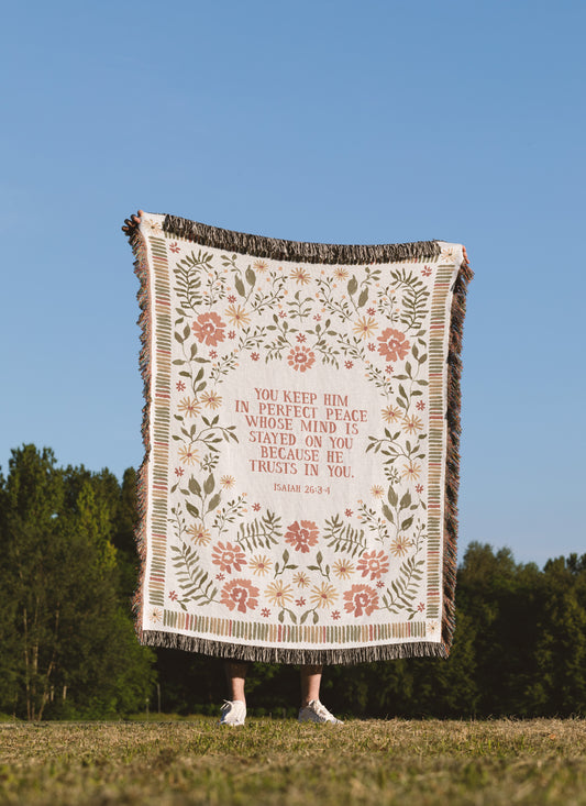 A person outdoors holding up a large cream-colored woven blanket with a floral pattern and a Bible verse printed in the center. The blanket has dark brown fringe on all sides.