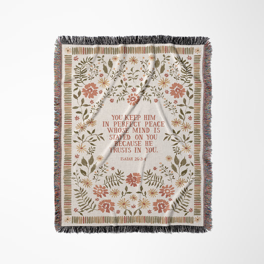 A rectangular woven throw blanket with fringe on all sides, featuring a central white arch motif surrounded by a floral border in muted pink, green, and beige tones. The center contains the printed text, 'YOU KEEP HIM IN PERFECT PEACE WHO'S MIND IS STAYED ON YOU BECAUSE HE TRUSTS IN YOU.
