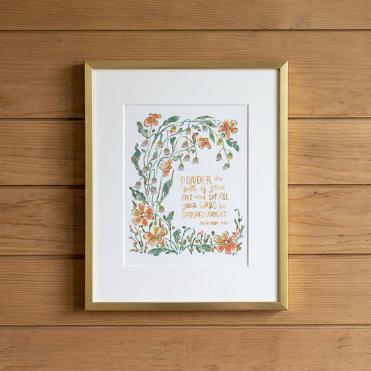 A framed art print featuring a botanical illustration with orange flowers and green leaves surrounding a handwritten quote, displayed on a light wood plank wall.