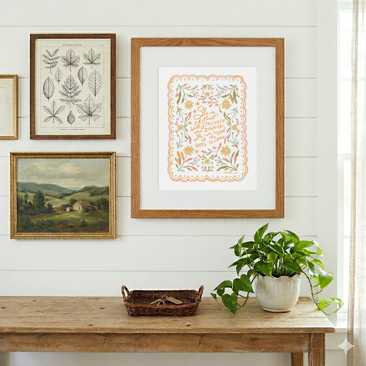 A framed art print featuring a colorful floral border and the words 'The Sweetest Thing' in orange script, displayed on a white shiplap wall above a rustic wooden table with a potted green plant.