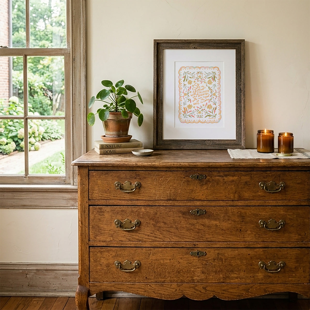 A framed botanical art print with the quote 'The best things in life are free' displayed on a rustic wooden dresser next to a window, with a potted plant and candles on top.