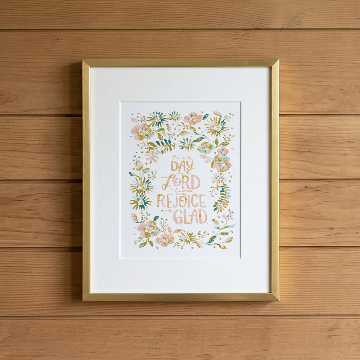 A framed art print featuring the hand-lettered quote 'DAY AND LORD AND REJOICE GLAD' surrounded by a watercolor floral wreath, displayed on a light wood plank wall.